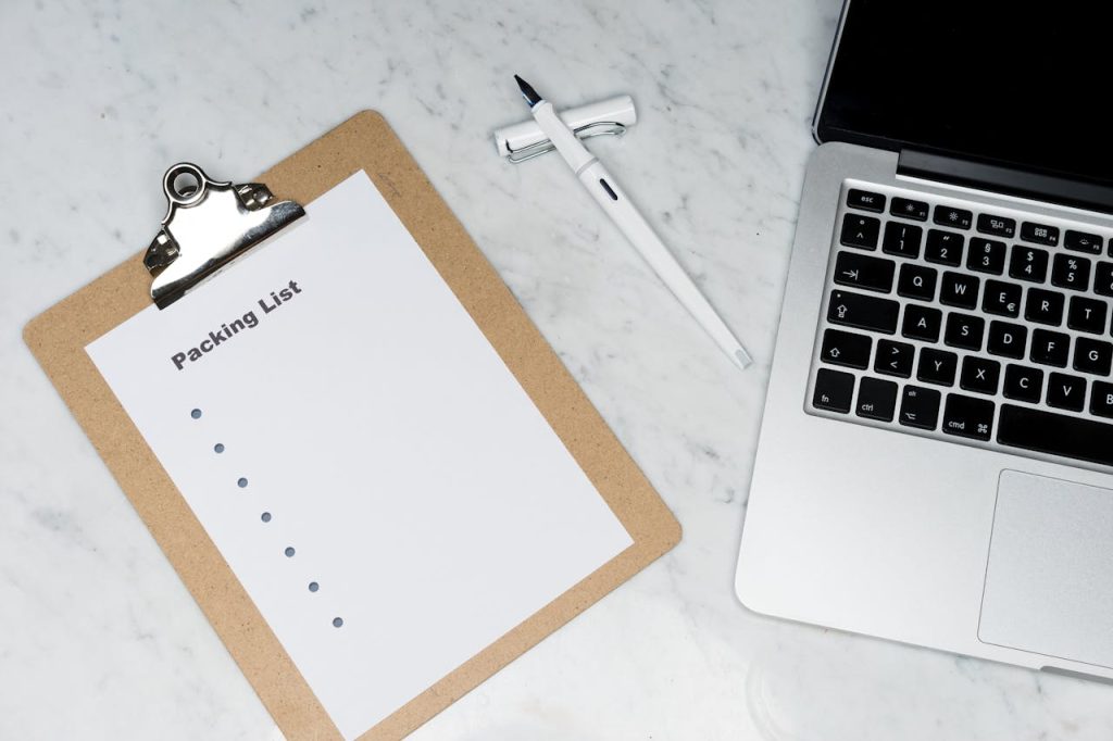 Flat lay of a packing list clipboard with a laptop and pen on a marble desk.
