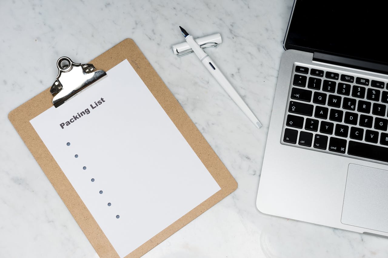 Flat lay of a packing list clipboard with a laptop and pen on a marble desk.